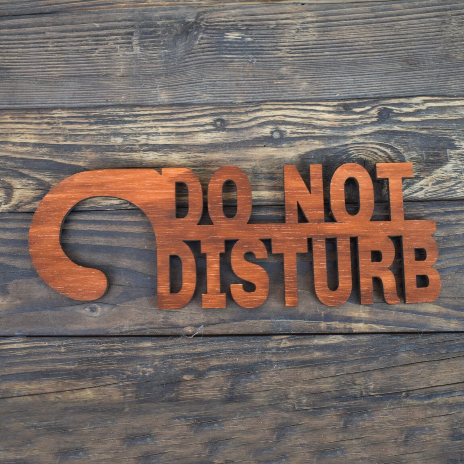 Ξύλινη Πινακίδα για Πόρτα Do not Disturb 6 This is a minimalist, laser-cut wooden door hanger used to signal privacy in a hotel or guestroom. The piece is crafted from light natural wood with a smooth, even grain. It has the classic door-hanger shape: a long vertical panel with a curved hook at the top that slips easily over a door handle. What makes this sign unique is that the message is not printed on the wood—the letters themselves are cut out as part of the sign. The text “DO NOT DISTURB” appears in large, bold capital letters that are carved completely through the wood, giving the sign an open, airy look. The letters are thick and sturdy, arranged in three stacked lines, with each word aligned vertically. Because the letters are cut out, the sign has a sculptural, three-dimensional feel, combining both function and design. The overall look is simple, modern, and elegant, making it ideal for boutique hotels, Airbnb stays, spas, or private rooms. Light brown