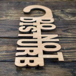 This is a minimalist, laser-cut wooden door hanger used to signal privacy in a hotel or guestroom. The piece is crafted from light natural wood with a smooth, even grain. It has the classic door-hanger shape: a long vertical panel with a curved hook at the top that slips easily over a door handle. What makes this sign unique is that the message is not printed on the wood—the letters themselves are cut out as part of the sign. The text “DO NOT DISTURB” appears in large, bold capital letters that are carved completely through the wood, giving the sign an open, airy look. The letters are thick and sturdy, arranged in three stacked lines, with each word aligned vertically. Because the letters are cut out, the sign has a sculptural, three-dimensional feel, combining both function and design. The overall look is simple, modern, and elegant, making it ideal for boutique hotels, Airbnb stays, spas, or private rooms.