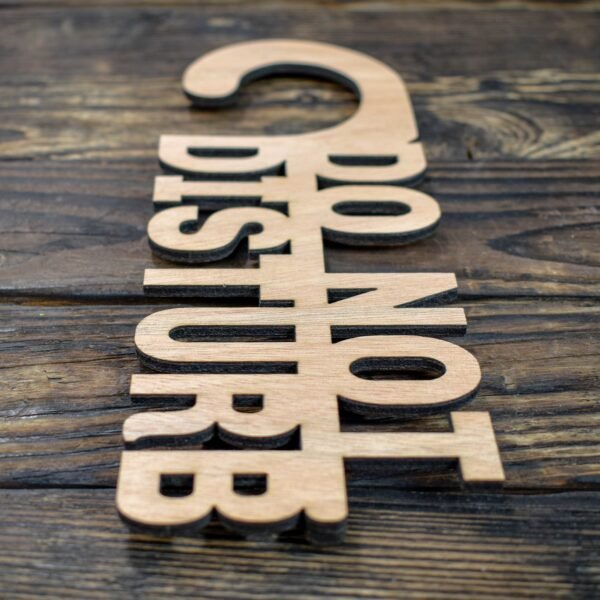 This is a minimalist, laser-cut wooden door hanger used to signal privacy in a hotel or guestroom. The piece is crafted from light natural wood with a smooth, even grain. It has the classic door-hanger shape: a long vertical panel with a curved hook at the top that slips easily over a door handle. What makes this sign unique is that the message is not printed on the wood—the letters themselves are cut out as part of the sign. The text “DO NOT DISTURB” appears in large, bold capital letters that are carved completely through the wood, giving the sign an open, airy look. The letters are thick and sturdy, arranged in three stacked lines, with each word aligned vertically. Because the letters are cut out, the sign has a sculptural, three-dimensional feel, combining both function and design. The overall look is simple, modern, and elegant, making it ideal for boutique hotels, Airbnb stays, spas, or private rooms.