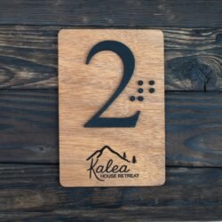 Πρόκειται για μία ορθογώνια ξύλινη πινακίδα, περίπου στο μέγεθος μιας παλάμης, με τις άκρες στρογγυλεμένες. Το ξύλο έχει φυσική, ζεστή απόχρωση — σαν καρυδιά — και η επιφάνειά του είναι λεία και απαλά γυαλισμένη, αλλά διατηρεί αισθητή την υφή των νερών του ξύλου. Στο κέντρο της πινακίδας υπάρχει ο αριθμός «2». Ο αριθμός δεν είναι τυπωμένος, αλλά ανασηκωμένος πάνω από το ξύλο — δηλαδή μπορείς να τον ψηλαφίσεις καθαρά. Είναι αρκετά μεγάλος, με ύψος περίπου όσο το μισό της πινακίδας. Το υλικό του είναι σκούρο, σχεδόν μαύρο, και η αφή του είναι λεία. Δίπλα στον αριθμό, προς τα δεξιά, υπάρχουν ανάγλυφες τελείες σε διάταξη Braille, ώστε κάποιος να μπορεί να διαβάσει τον αριθμό «2» με την αφή. Οι τελείες είναι μικρές και εξίσου ανασηκωμένες. Στο κάτω μέρος της πινακίδας, υπάρχει χαραγμένο ένα μικρό σχέδιο: ένα βουνό με δύο μικρά δέντρα και από κάτω μια λέξη γραμμένη με καλλιγραφικά γράμματα. Το σχέδιο είναι χαραγμένο μέσα στο ξύλο, οπότε στην αφή είναι ελαφρώς πιο βαθύ από την επιφάνεια. Δεν εξέχει.