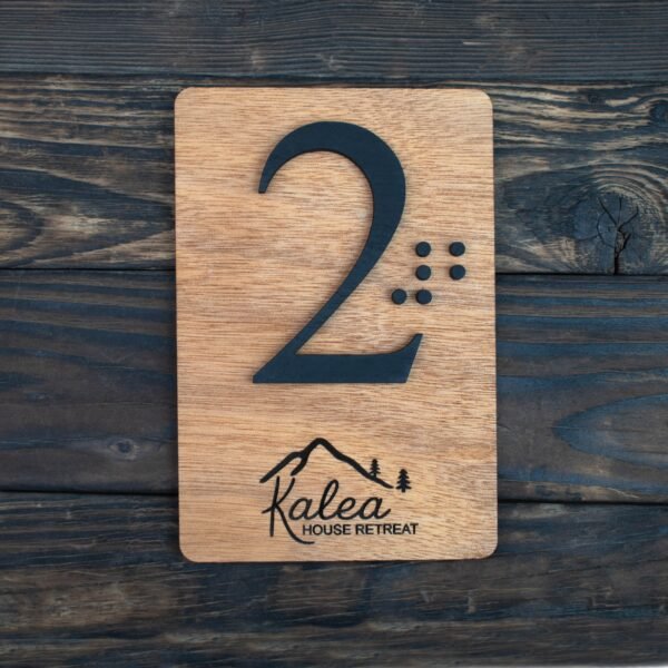 Πρόκειται για μία ορθογώνια ξύλινη πινακίδα, περίπου στο μέγεθος μιας παλάμης, με τις άκρες στρογγυλεμένες. Το ξύλο έχει φυσική, ζεστή απόχρωση — σαν καρυδιά — και η επιφάνειά του είναι λεία και απαλά γυαλισμένη, αλλά διατηρεί αισθητή την υφή των νερών του ξύλου. Στο κέντρο της πινακίδας υπάρχει ο αριθμός «2». Ο αριθμός δεν είναι τυπωμένος, αλλά ανασηκωμένος πάνω από το ξύλο — δηλαδή μπορείς να τον ψηλαφίσεις καθαρά. Είναι αρκετά μεγάλος, με ύψος περίπου όσο το μισό της πινακίδας. Το υλικό του είναι σκούρο, σχεδόν μαύρο, και η αφή του είναι λεία. Δίπλα στον αριθμό, προς τα δεξιά, υπάρχουν ανάγλυφες τελείες σε διάταξη Braille, ώστε κάποιος να μπορεί να διαβάσει τον αριθμό «2» με την αφή. Οι τελείες είναι μικρές και εξίσου ανασηκωμένες. Στο κάτω μέρος της πινακίδας, υπάρχει χαραγμένο ένα μικρό σχέδιο: ένα βουνό με δύο μικρά δέντρα και από κάτω μια λέξη γραμμένη με καλλιγραφικά γράμματα. Το σχέδιο είναι χαραγμένο μέσα στο ξύλο, οπότε στην αφή είναι ελαφρώς πιο βαθύ από την επιφάνεια. Δεν εξέχει.