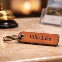 Δερμάτινο Μπρελόκ για Ξενοδοχείο, Πανσιόν, Ενοικιαζόμενα Δωμάτια και Airnbn 3 Δερμάτινο μπρελόκ με χάραξη «Villa Lisa» πάνω σε γυαλιστερό πάγκο υποδοχής ξενοδοχείου, με μπρούτζινο κουδούνι και φυλλάδια θολά στο φόντο υπό ζεστό φωτισμό.
