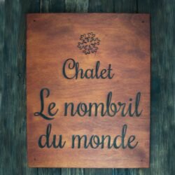 Ξύλινη Πινακίδα με Όνομα Καταλύματος 50Χ30cm 6 This is a rectangular wooden sign with a smooth, warm reddish-brown finish. The wood grain is visible, giving the piece a natural and rustic character, suitable for a mountain chalet or cozy holiday home. At the top center of the sign, there is a carved snowflake symbol. The snowflake has delicate, symmetrical branches, adding a wintery, alpine touch to the design. Beneath the snowflake, the sign features engraved text in French. The lettering is done in a flowing cursive script, elegant and easy to read by touch. The text is centered and arranged across three lines: “Chalet Le nombril du monde” The engraved letters are darkened, creating a clear contrast against the lighter wood surface. The overall style is simple, welcoming, and charming—perfect for identifying a chalet with personality and warmth.