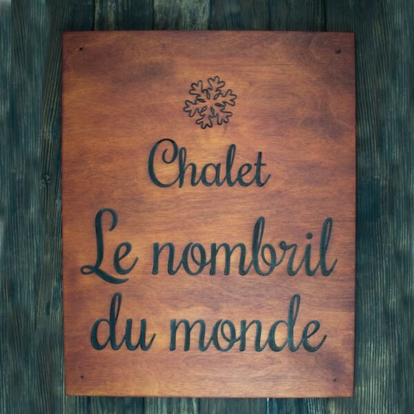 This is a rectangular wooden sign with a smooth, warm reddish-brown finish. The wood grain is visible, giving the piece a natural and rustic character, suitable for a mountain chalet or cozy holiday home. At the top center of the sign, there is a carved snowflake symbol. The snowflake has delicate, symmetrical branches, adding a wintery, alpine touch to the design. Beneath the snowflake, the sign features engraved text in French. The lettering is done in a flowing cursive script, elegant and easy to read by touch. The text is centered and arranged across three lines: “Chalet Le nombril du monde” The engraved letters are darkened, creating a clear contrast against the lighter wood surface. The overall style is simple, welcoming, and charming—perfect for identifying a chalet with personality and warmth.
