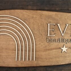 Χειροποίητη Ξύλινη Πινακίδα Σήμανσης Καταλύματος 4 This is an elegant, handcrafted wooden sign designed for a boutique hotel or spa. The sign has an elongated oval shape with softly rounded edges, made from warm, medium-brown stained wood. The surface is smooth and the wood grain is visible, giving it a natural, refined look. On the left side of the sign, there is a raised decorative motif made of thin, curved wooden lines. These lines form a stylized, minimalist rainbow composed of seven parallel arches. The arches are evenly spaced and follow the same rounded shape, creating a clean and calming visual pattern. On the right side, the hotel’s name “EVEXIA” appears in large, raised capital letters. The font is elegant and classic, with thin lines and gentle curves. Beneath it, in smaller raised letters, is the phrase “boutique hotel & spa.” Below the text, four raised wooden stars are arranged in a horizontal row, representing a four-star accommodation rating. The overall look of the sign is sophisticated and modern, combining natural wood with clean design elements. It’s meant to convey quality, warmth, and a welcoming atmosphere.