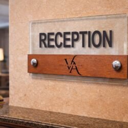 Πινακίδα Plexiglass και Ξύλο Με Λογότυπο 3 Επίτοιχη πινακίδα Reception από διάφανο ακρυλικό και ξύλο κόντρα πλακέ με ανάγλυφα μαύρα γράμματα και λογότυπο σε τοίχο υποδοχής.