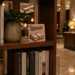 Χειροποίητη επιτραπέζια ξύλινη πινακίδα σήμανσης με τη λέξη "Library" και ένα τρισδιάστατο (3D) επικολλημένο σχέδιο ανοιχτού βιβλίου, τοποθετημένη σε ράφι βιβλιοθήκης ξενοδοχείου.