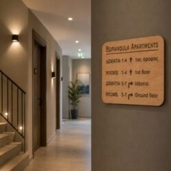 Χειροποίητη ξύλινη πινακίδα κατεύθυνσης τοποθετημένη σε τοίχο μοντέρνου διαδρόμου ξενοδοχείου, με χαραγμένες οδηγίες δωματίων στα ελληνικά και αγγλικά.